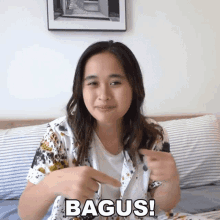 a woman is sitting on a bed pointing at the camera with the word bagus written on her chest .