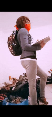 a woman wearing a mask and a backpack is standing in a messy room holding a book