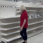 a woman in a red shirt is standing in front of a row of empty shelves in a store .