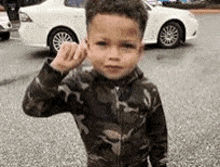 a young boy in a camouflage jacket is standing in a parking lot with a white car in the background .