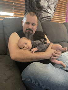 a man with a beard is holding a baby on a couch with a picture of a cat behind him