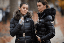 two women standing next to each other wearing black jackets with the letter h on the front