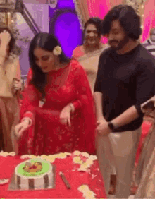 a woman in a red dress is cutting a cake with a knife while a man stands behind her .