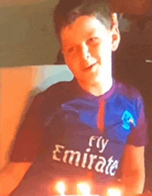 a young boy wearing a fly emirates shirt is sitting in front of a cake with candles .