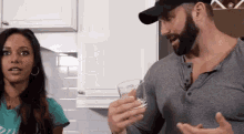 a man with a beard is holding a glass of water while talking to a woman in a kitchen .