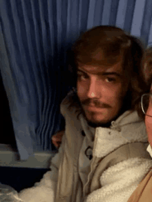 a man with long hair and a mustache is sitting next to another man on a plane .