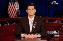 a man in a suit and tie is sitting at a desk with his hands folded in front of a screen that says live on it