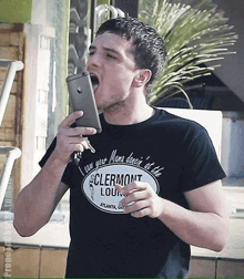 a man wearing a black shirt that says clermont lounge holds a cell phone in his mouth