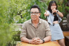 a man with glasses sits at a table with his hands folded and a woman behind him pointing