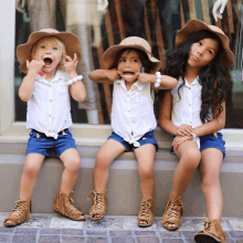 three little girls wearing hats and shorts are sitting on a wall making funny faces