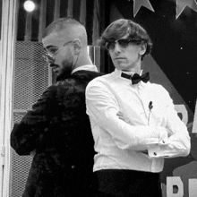 a black and white photo of two men standing back to back . one of the men is wearing sunglasses and a bow tie .