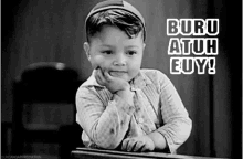 a black and white photo of a young boy sitting at a desk with his hand on his chin and the words buru atuh cuy .