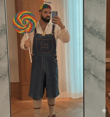 a man in overalls is holding a lollipop and taking a selfie in a mirror