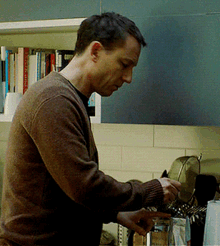 a man in a brown sweater is cooking in a kitchen with a bookshelf behind him that has cook 's book on it