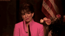 a woman in a pink suit is smiling and says `` you 're welcome '' in front of an american flag .