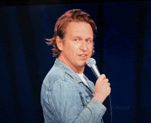 a man in a denim jacket holds a microphone in front of a blue curtain