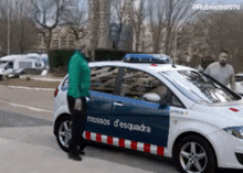 a man in a green jacket is standing next to a police car that says mossos d' esquadra on the side