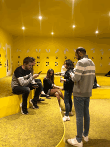 a group of people in a room with a yellow wall that has the letters k m n o p s and t on it