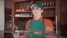 a man wearing a green apron and an orange shirt is standing in front of a glass display case .