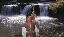 a man is holding a sword in front of a waterfall in a river .