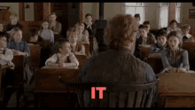 a man sits in a chair in front of a classroom full of children with the word it in red