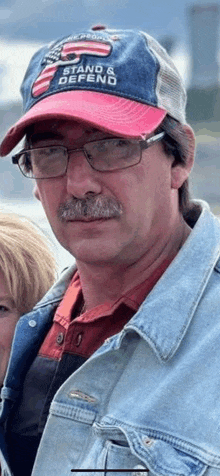 a man with a mustache and glasses is wearing a red white and blue hat and a denim jacket .