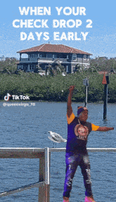 a man in a purple shirt stands on a dock near a body of water with a house in the background