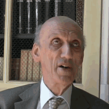 a bald man in a suit and tie stands in front of a bookshelf with a few books on it