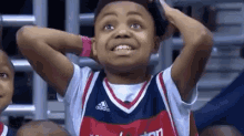 a young boy in a washington wizards jersey is sitting in the stands with his hands on his head .