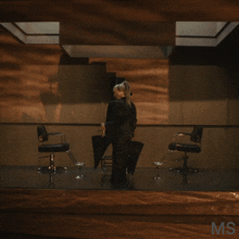 a woman in a black suit is dancing in a room with chairs and a sign that says ms on it
