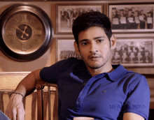 a man in a blue shirt is leaning on a chair in front of a wall clock that says xii on it