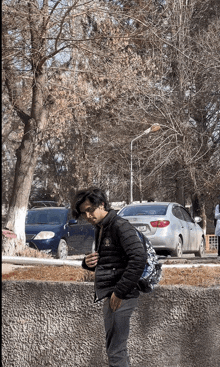 a man in a black jacket with a backpack stands in front of a car with a license plate that starts with the letter t