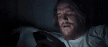 a man with a beard is laying in bed looking at his phone in the dark .