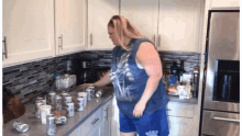 a woman in a blue tank top is standing in a kitchen with a lot of cans on the counter