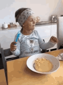 a little girl is sitting at a table with a bowl of food and a spoon in her hand .