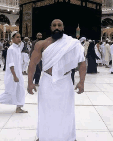 a man with a beard and a white robe is standing in front of a kaaba .