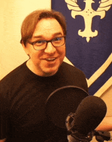 a man wearing glasses and a black shirt stands in front of a microphone in front of a flag with the letter i on it