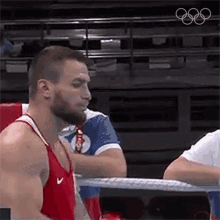 a man in a red tank top is standing in a boxing ring with his hand on his chest .