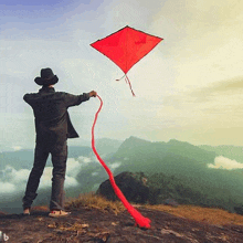 a man in a hat is flying a red kite on top of a hill .