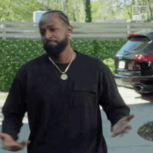 a man with a beard wearing a black shirt and a gold necklace is standing in front of a black car .