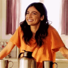 a woman in an orange shirt is smiling while sitting at a table with coffee mugs and a pitcher .