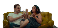 a man and a woman sit on a yellow couch eating ice cream cones