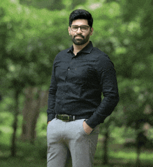 a man wearing glasses and a black shirt stands in front of a green forest