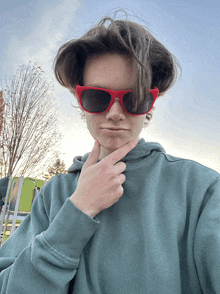 a person wearing a green sweatshirt and red sunglasses holds their hand to their chin