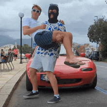 a man carrying another man in front of a red car with a license plate that says k3