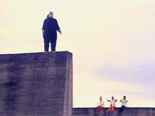 a man in a mask is standing on top of a concrete wall with a group of people sitting below him