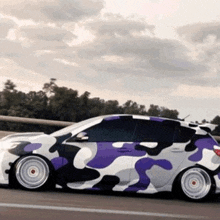 a white car with a purple and black camouflage wrap is driving down a road .