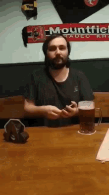 a man with a beard is sitting at a table with a mug of beer and a mountfield scarf on the wall behind him