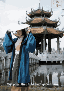 a woman wearing a blue coat and a white hat stands in front of a pagoda and a body of water