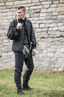 a man in a black jacket and pants is holding a sword in front of a stone wall .